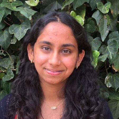 A person with black sweater and red shirt standing in front of leafy background.