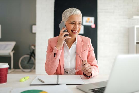 A woman with gray hair talking on the phone at a desk.