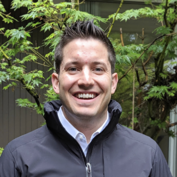 A person smiling outdoors with leafy background.