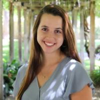 A person smiling outdoors with a greenery background.