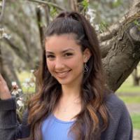 Person smiling outdoor with long, wavy hair and trees in the background.