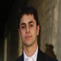 A man in a dark suit stands near a brick wall.