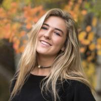 A person smiling in an outdoor setting with autumn leaves in the background.