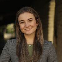 A person smiling outdoors, wearing a gray blazer.