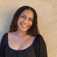 A person smiling while wearing a black top standing against a beige wall.