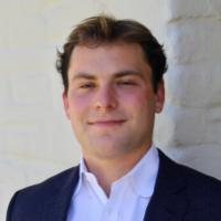 A person in a suit standing against a light-colored wall.