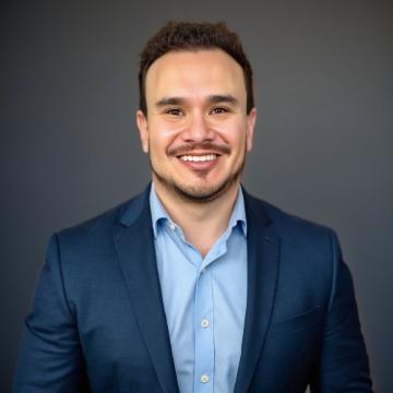 A photo of Guadalupe Hayes-Mota, smiling into the camera. He is wearing a light blue shirt with navy blazer.
