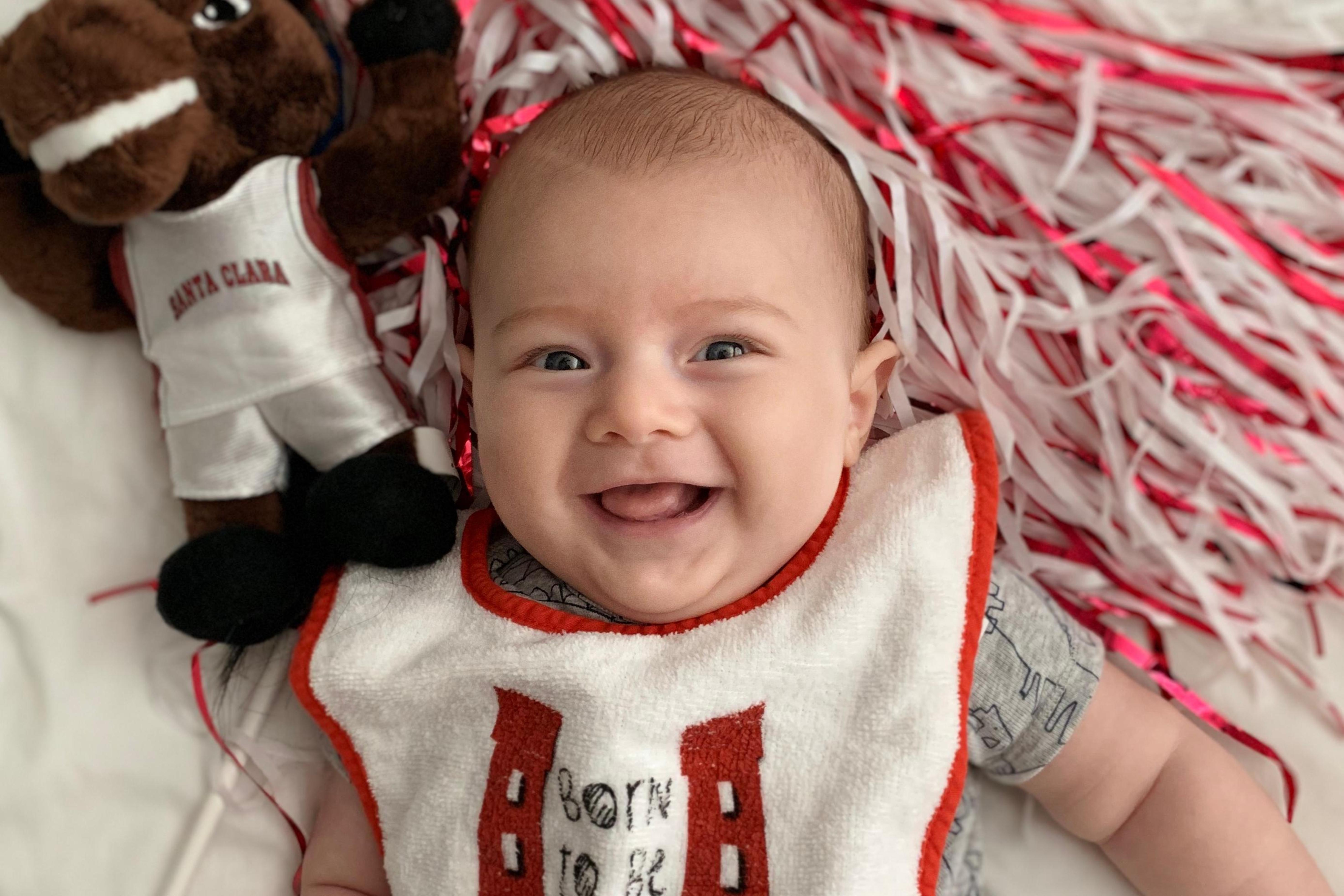 A smiling baby wearing a bib with a stuffed toy nearby.