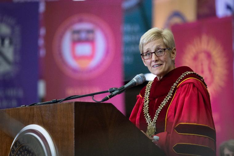 A person speaking at a podium, dressed in academic regalia, with colorful banners in the background.