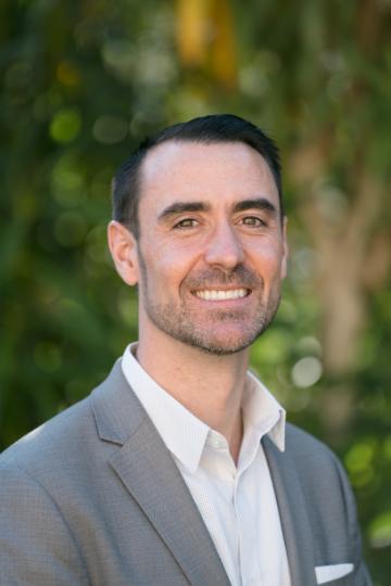 A man smiling in a suit against a green leafy background.