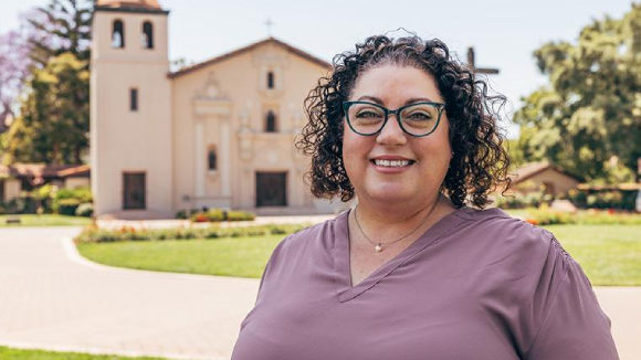 A smiling woman in glasses stands in front of Santa Clara University's Mission Church.