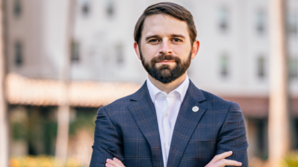 A man in a suit with his arms crossed in front of Nobili Hall at Santa Clara University