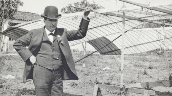 A black and white photo from the early 1900s of a man in a bowler hat and suit standing next to a glider