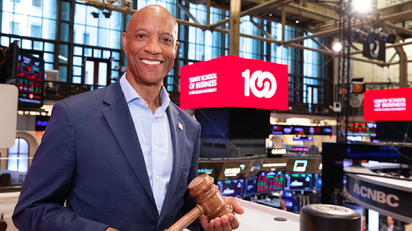 A smiling man holding a gavel at the New York Stock Exchange. Behind him is a red screen with white text reading 