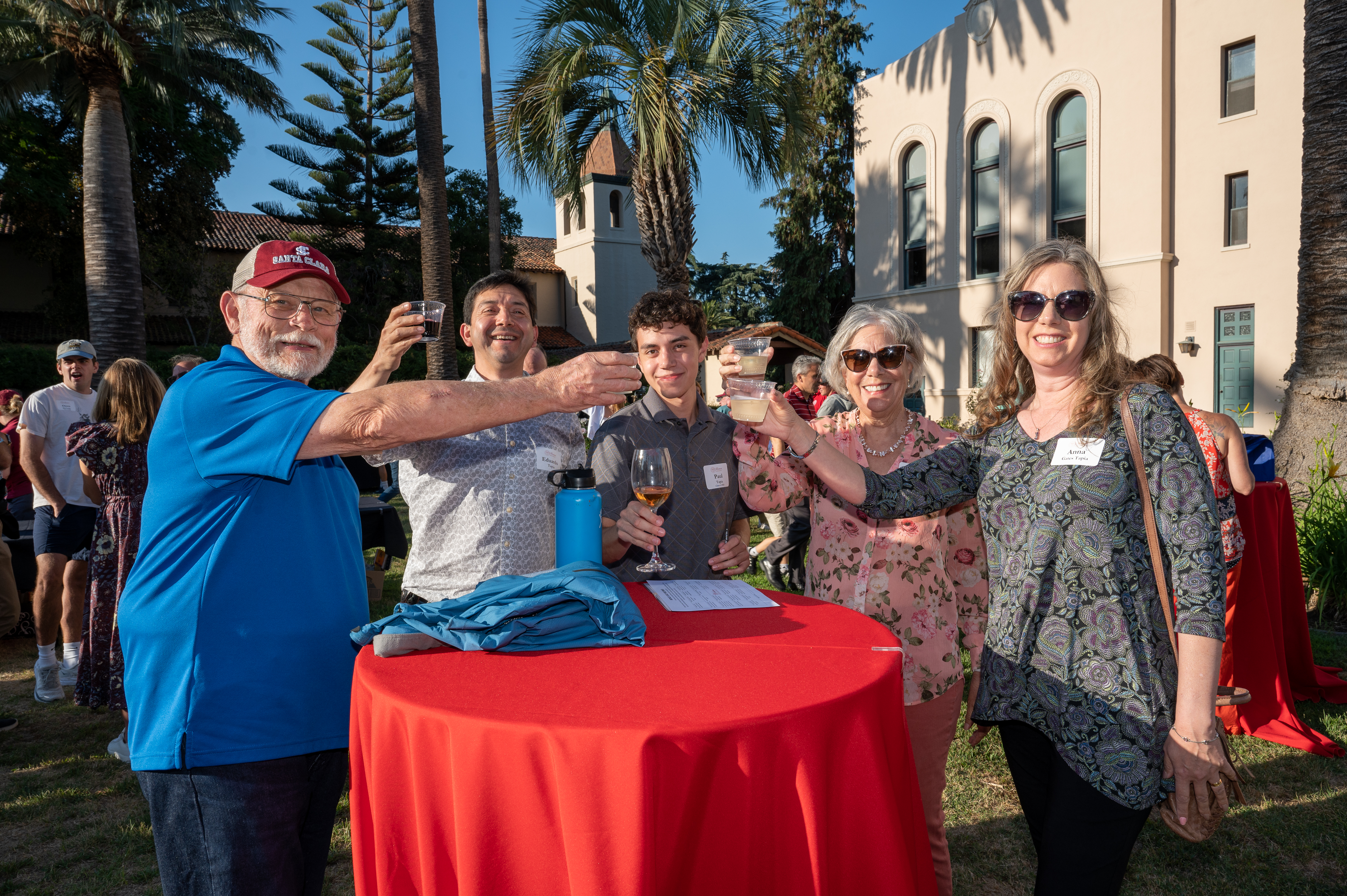 Group of people raising glasses at an outdoor event near a red table.