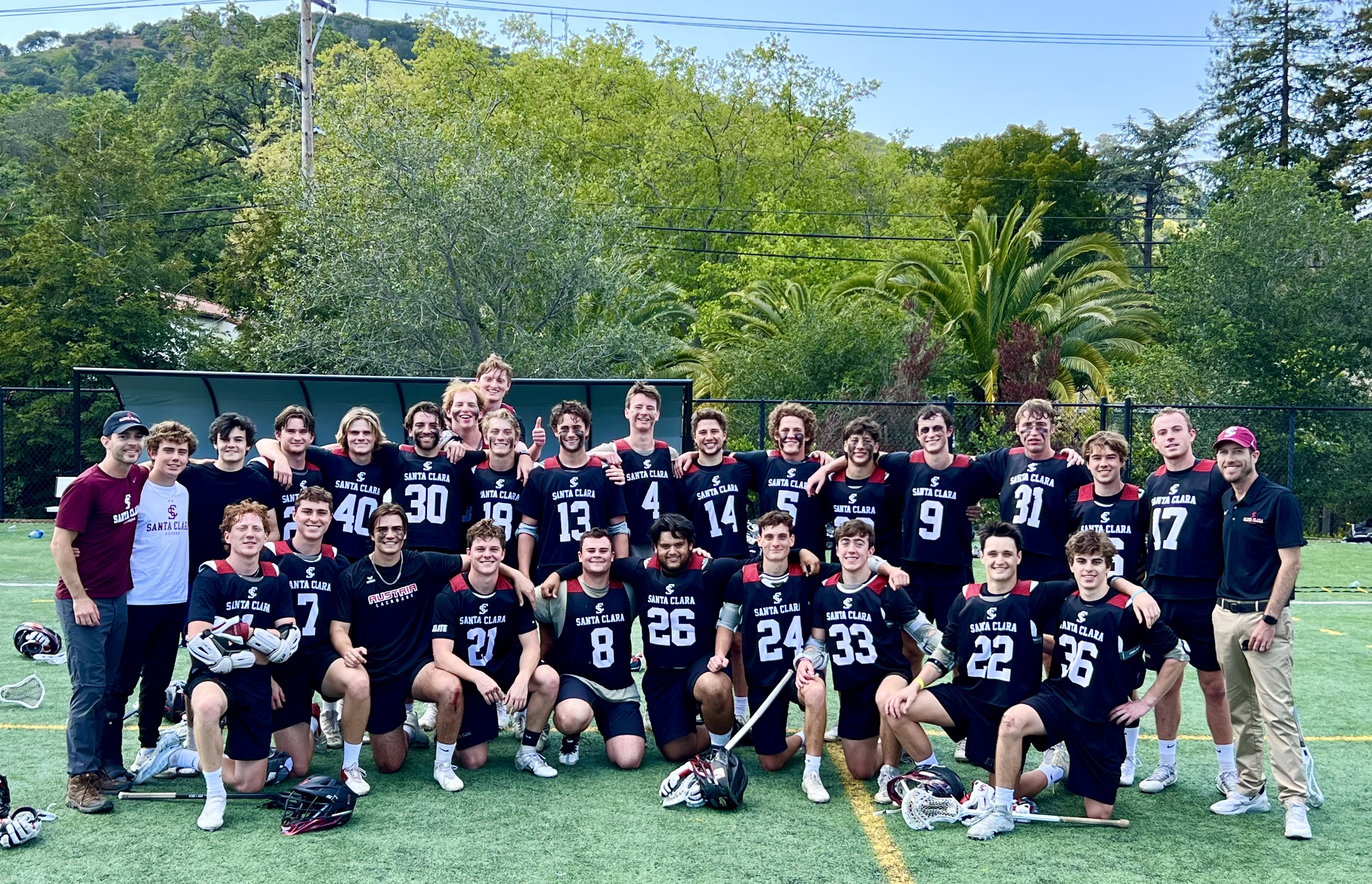 Men's lacrosse team posing on field, surrounded by trees.
