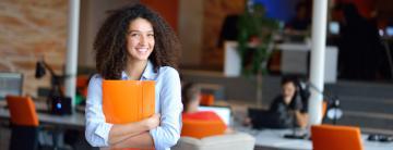 A woman holding orange folders, smiling in a modern office setting.