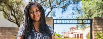 Smiling person outdoors with trees, brick wall, and building in the background.