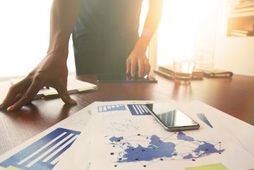 Person reviewing charts and documents on a desk with a pen and glasses.