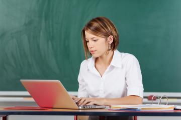 A teacher working on a laptop in front of a blackboard.