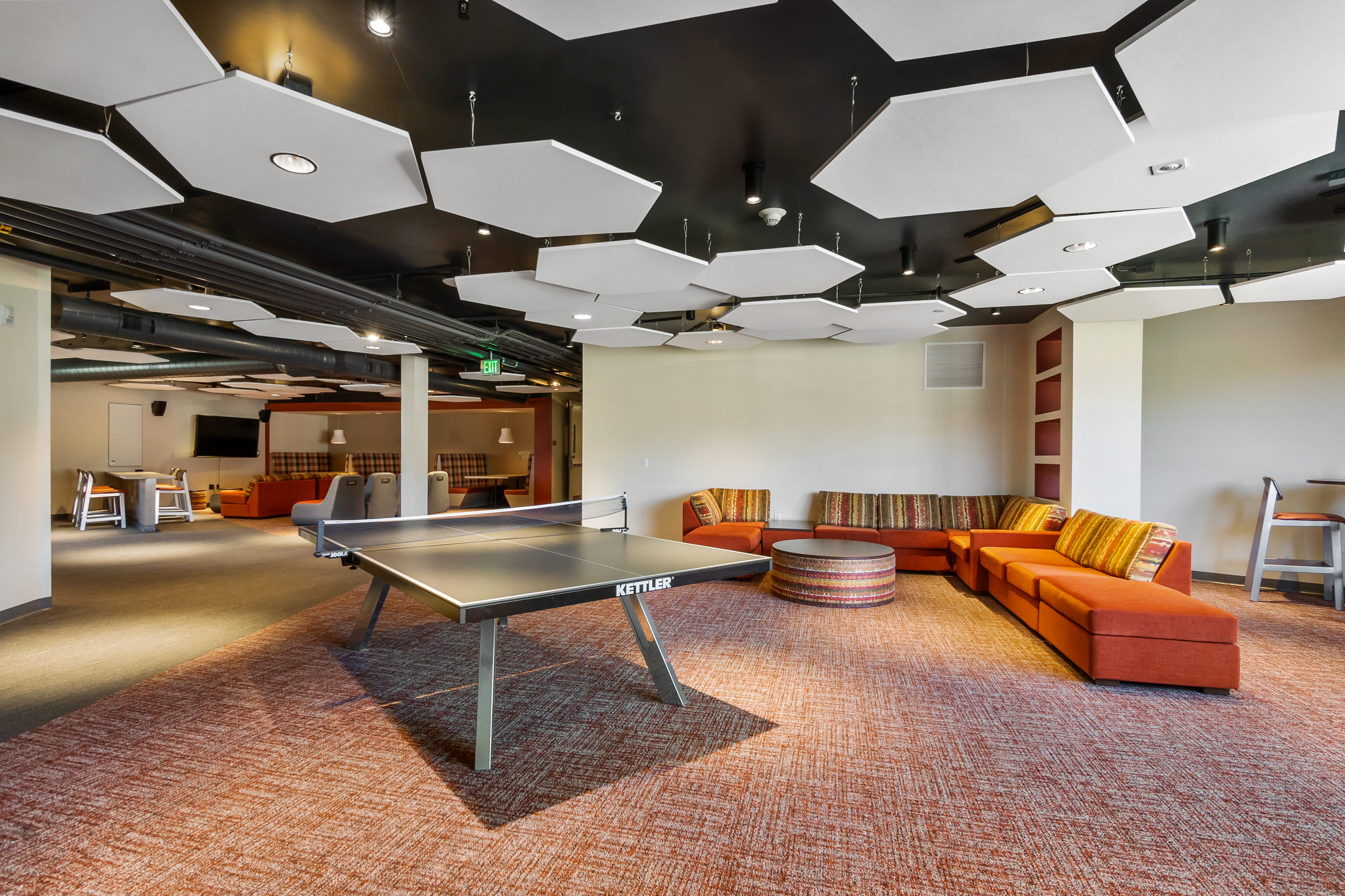Modern lounge area with hexagonal ceiling panels, orange couches, and wooden furniture.