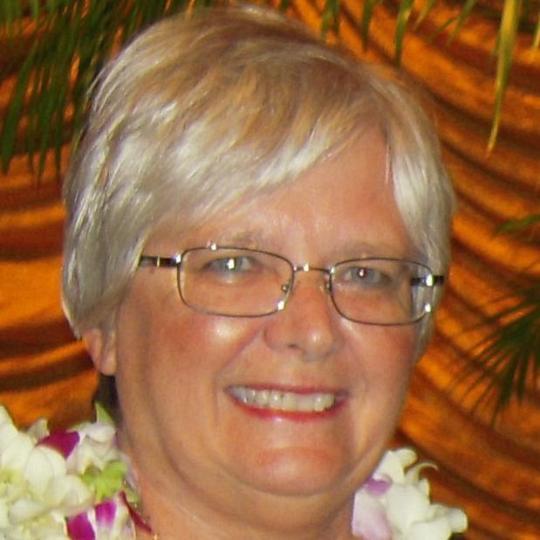 Alt text: Person wearing eyeglasses and leis, smiling in front of a patterned background.