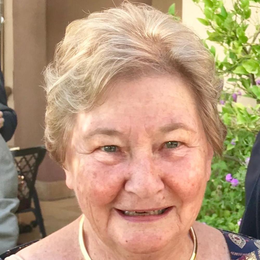A woman smiling, wearing a patterned blouse and necklace, standing outdoors.