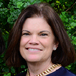 Woman in blue dress with necklace standing in front of greenery.
