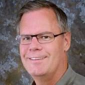 A person wearing glasses and smiling against a dark, textured background.