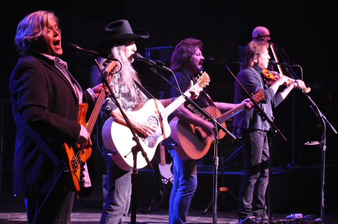 Four band members of The Doobie Brothers performing on stage with guitars and microphones.