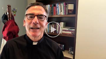 Man in clerical attire speaking indoors in front of a bookshelf.