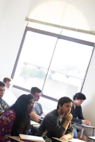 Students are seated at desks in a classroom with a large window.