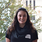 A person with long hair standing outdoors, trees in the background.