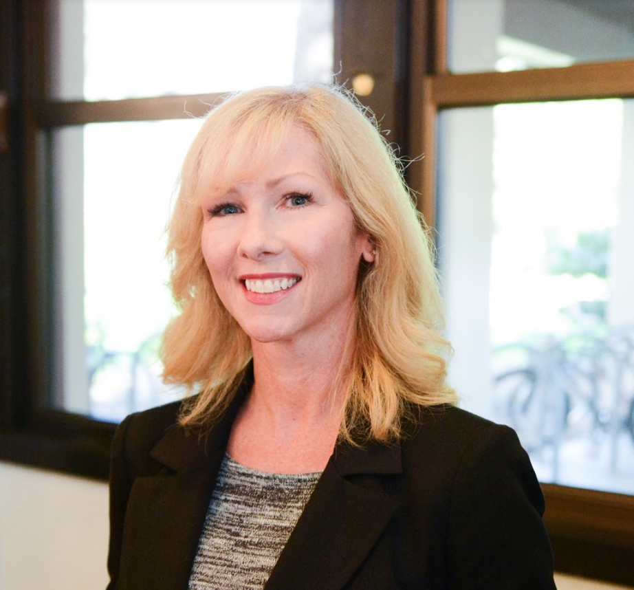 A woman smiling, standing in front of windows.