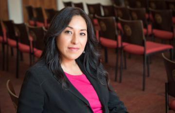 A woman in a black blazer sits in a room with chairs.