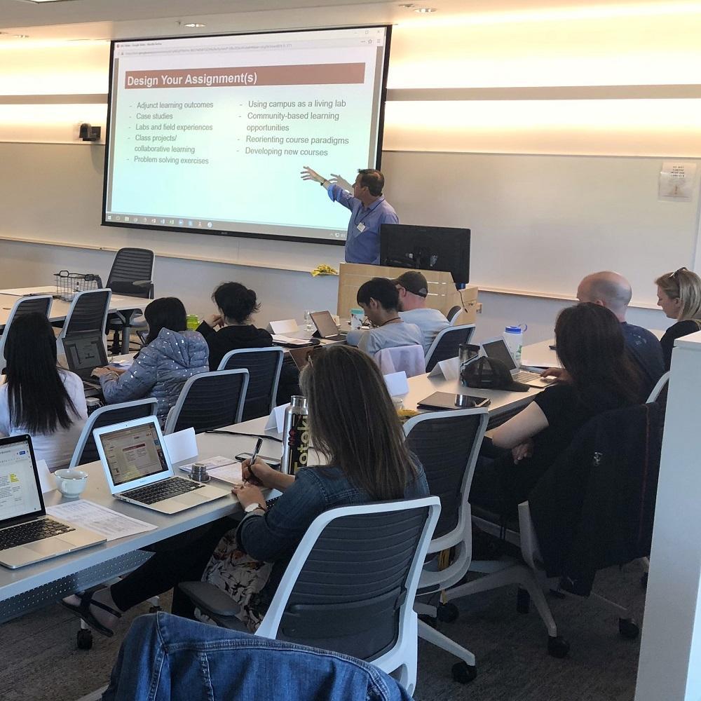 A teacher pointing to a projector in a room of seated teachers with their laptops on tables in front of them