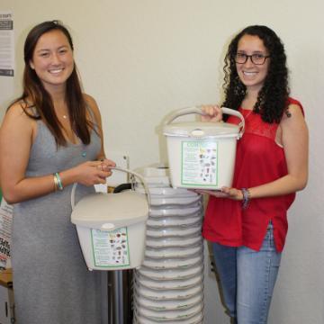 Two women holding compost bins in a room titled 