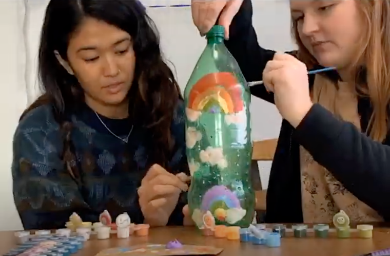 Two people painting an upcycled bottle with colorful designs.
