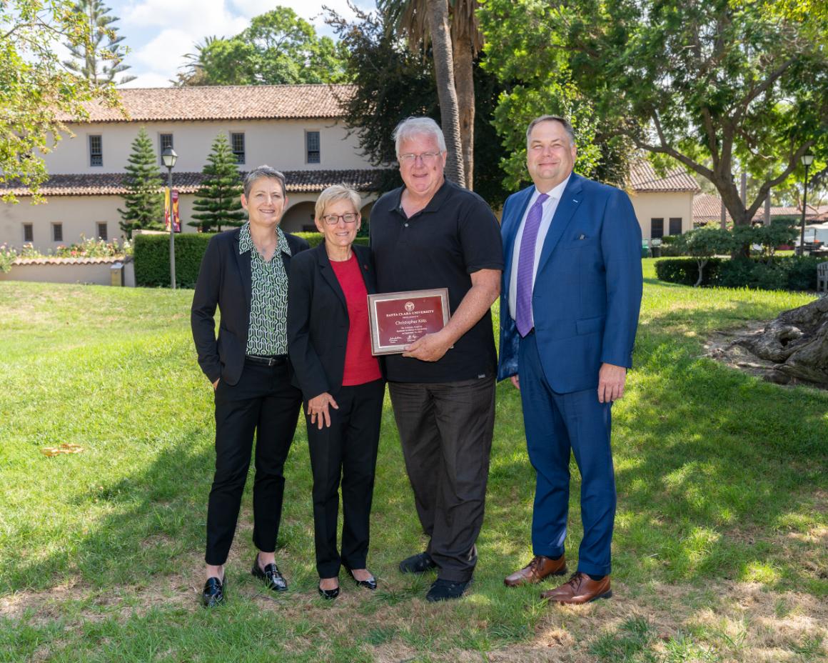 Four people standing outdoors, one holding an award plaque titled 