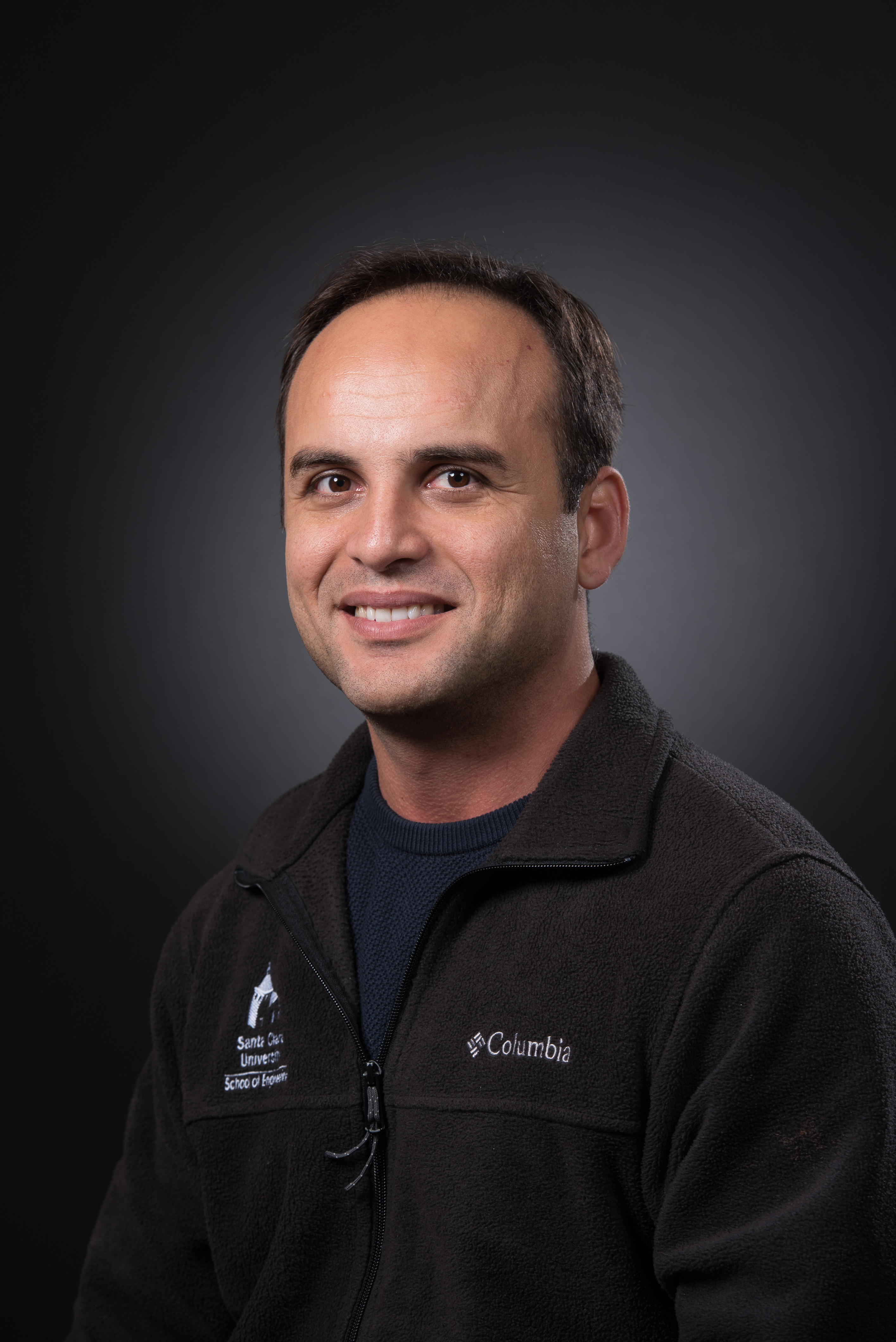 A person wearing a dark fleece jacket, smiling against a dark background.