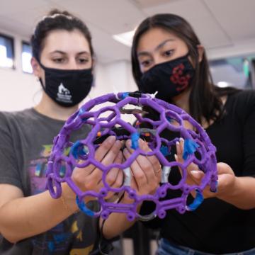 Two people examining a purple molecular model while wearing masks.