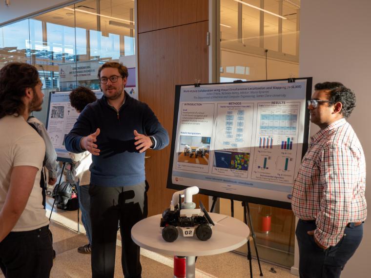 Two male students speaking with a male guest with a poster board behind them and a small robot on wheels on top of a table in front of them.