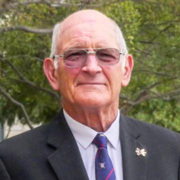 A man wearing glasses, a suit, and a tie, with trees in the background.