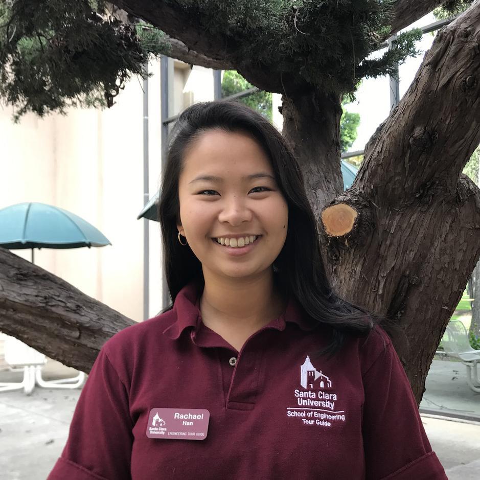 Person in maroon shirt with name tag, smiling outdoors, near a large tree.